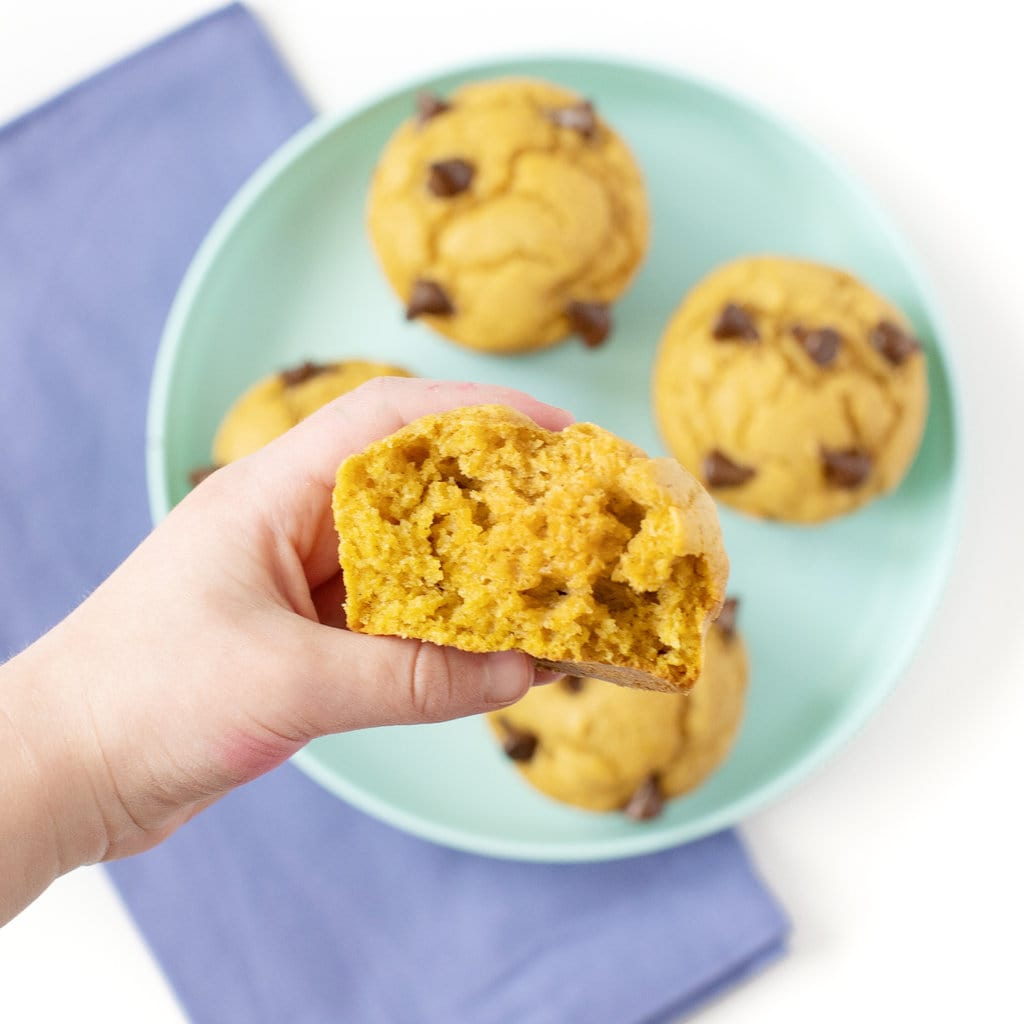 Teal kids plate with 4 sweet potato muffins and a dark blue napkin with a kids hand holding a torn in half mufifn.