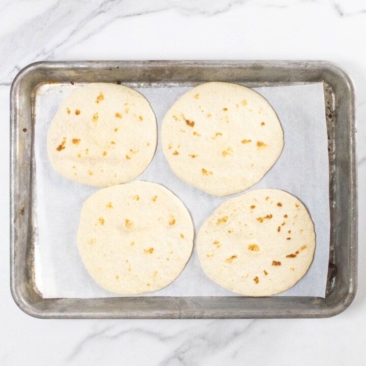 Baking sheet on a marble counter with four flour tortillas on a piece of parchment paper.