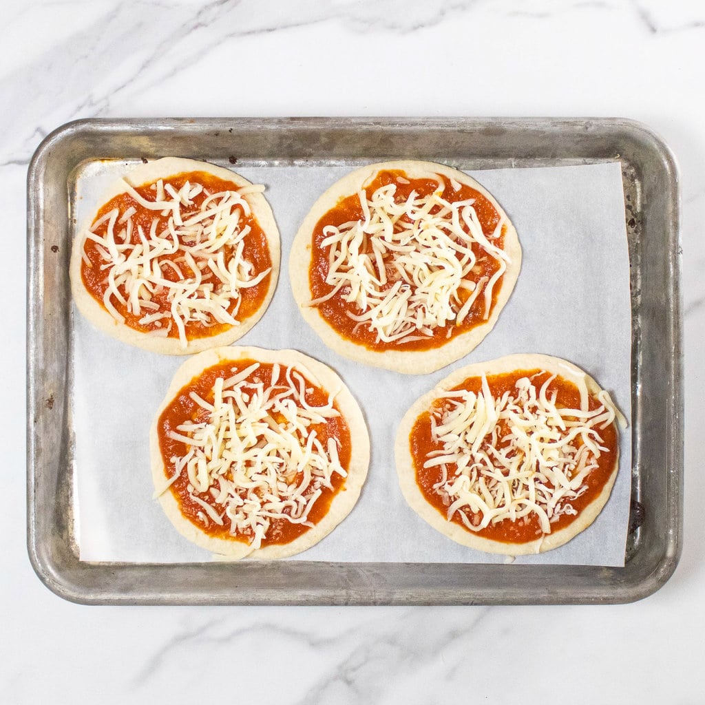 Baking sheet on a marble counter with four tortilla pizzas that are mini size with sau