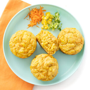 Three veggie muffins on a plate with one torn and a half with piles of carrot, zucchini and corn on a blue kids plate with a orange napkin against a white background.