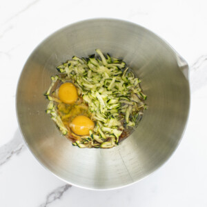 Silver mixing bowl on a marble counter filled with zucchini and other wet ingredients.