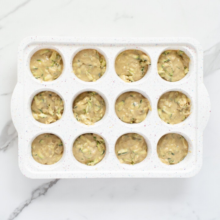 A white muffin pan on a white counter full of zucchini muffin dough.