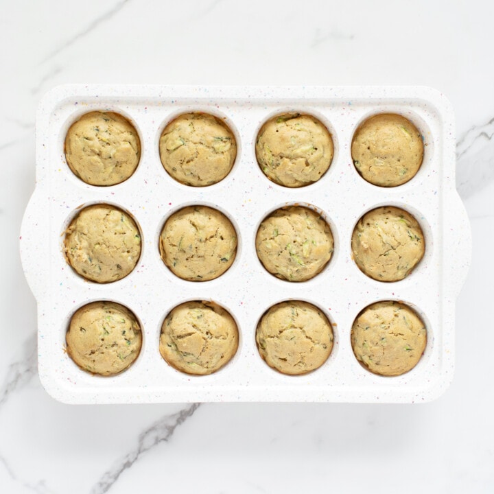 A white muffin tin on a white kitchen counter full of cooked zucchini muffins.