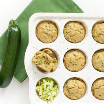 White muffin tin with cooked zucchini muffins on a green napkin with a zucchini sitting next to it and shredded zucchini in one of the muffin rings.