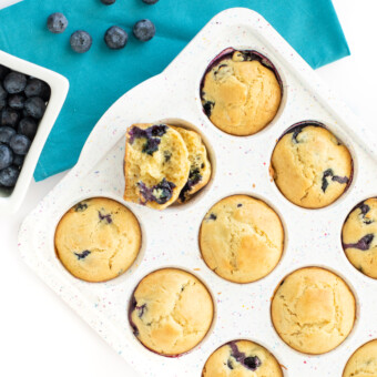 A speckled white muffin tray full with cooked blueberry muffins against away background with blue tablecloth and scatter blueberries around it.