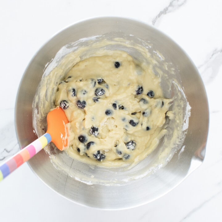Silver mixing bowl with blueberry muffin batter and a colorful spoon inside.
