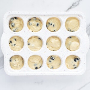 A white muffin pan on a white marble countertop with blueberry muffin batter.