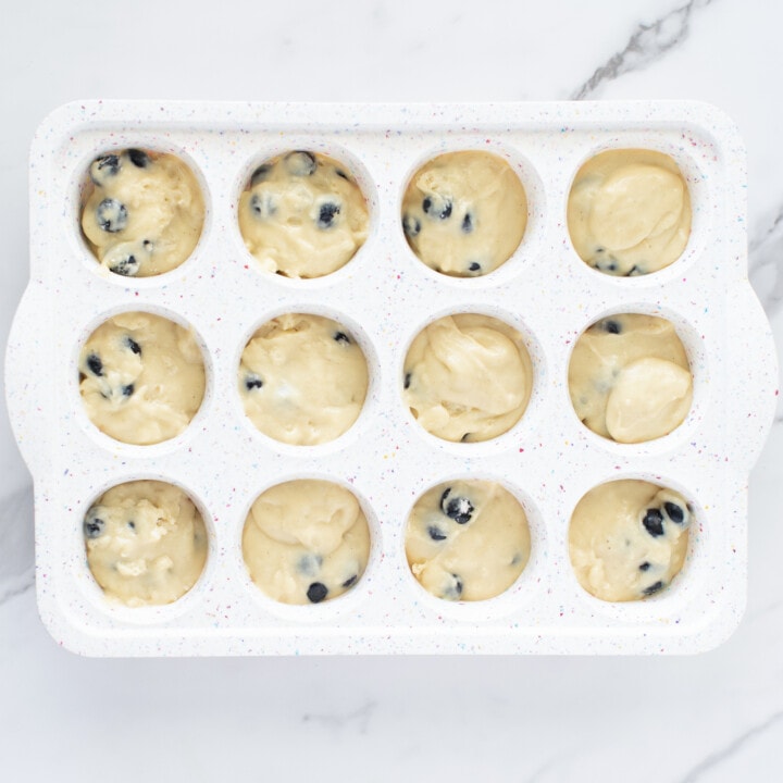 A white muffin pan on a white marble countertop with blueberry muffin batter.