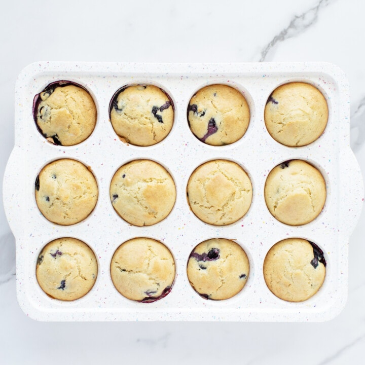 A white muffin tin on a white marble counter with baked blueberry muffins.