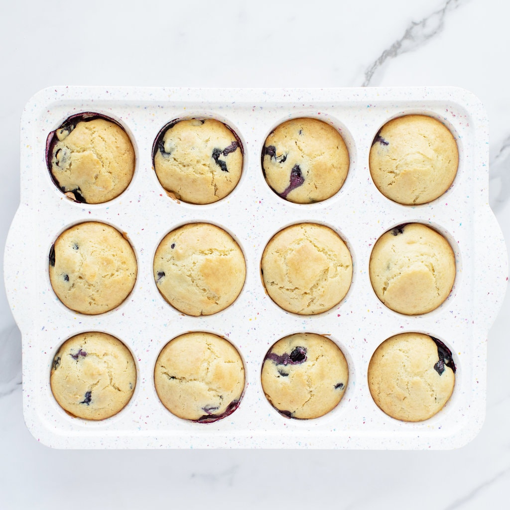 A white muffin tin on a white marble counter with baked blueberry muffins.