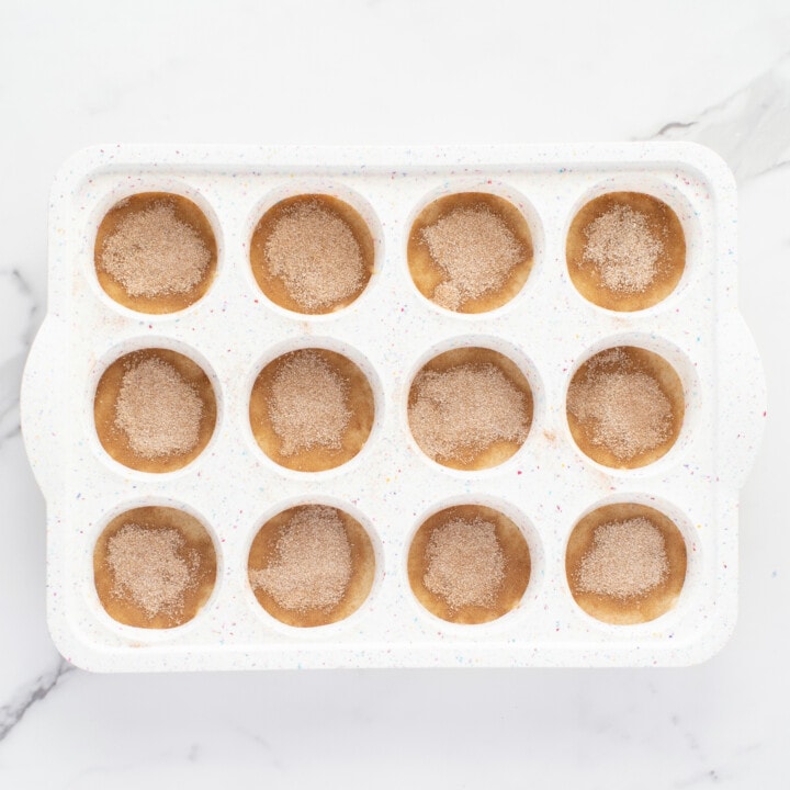 A white muffin tin with cinnamon sugar muffin ready to be cooked against a white marble background.