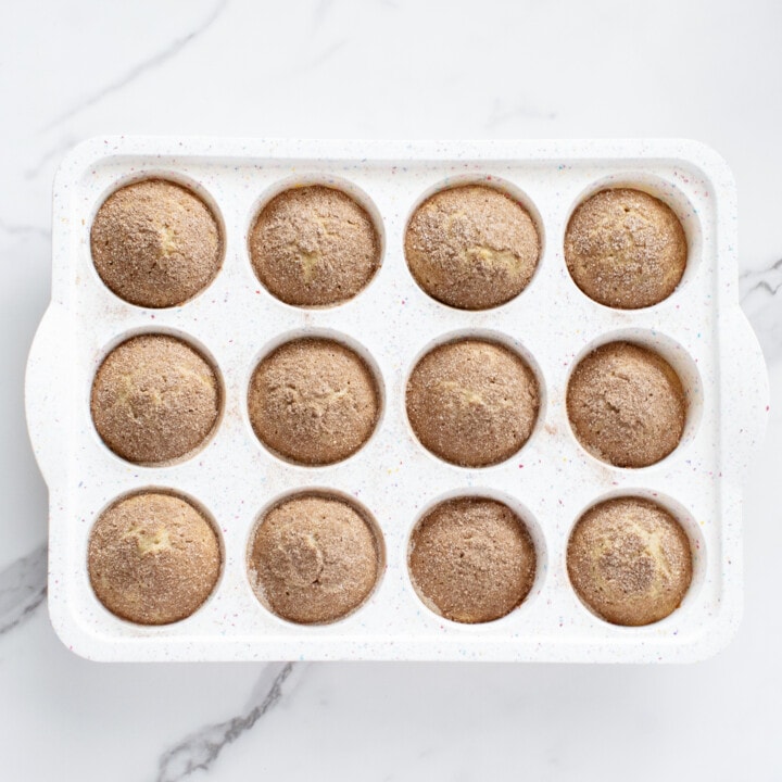 A white muffin tin filled with baked cinnamon muffins against a white marble countertop. 