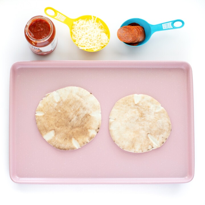 A pink baking sheet with ingredients for a kids pita pizza.