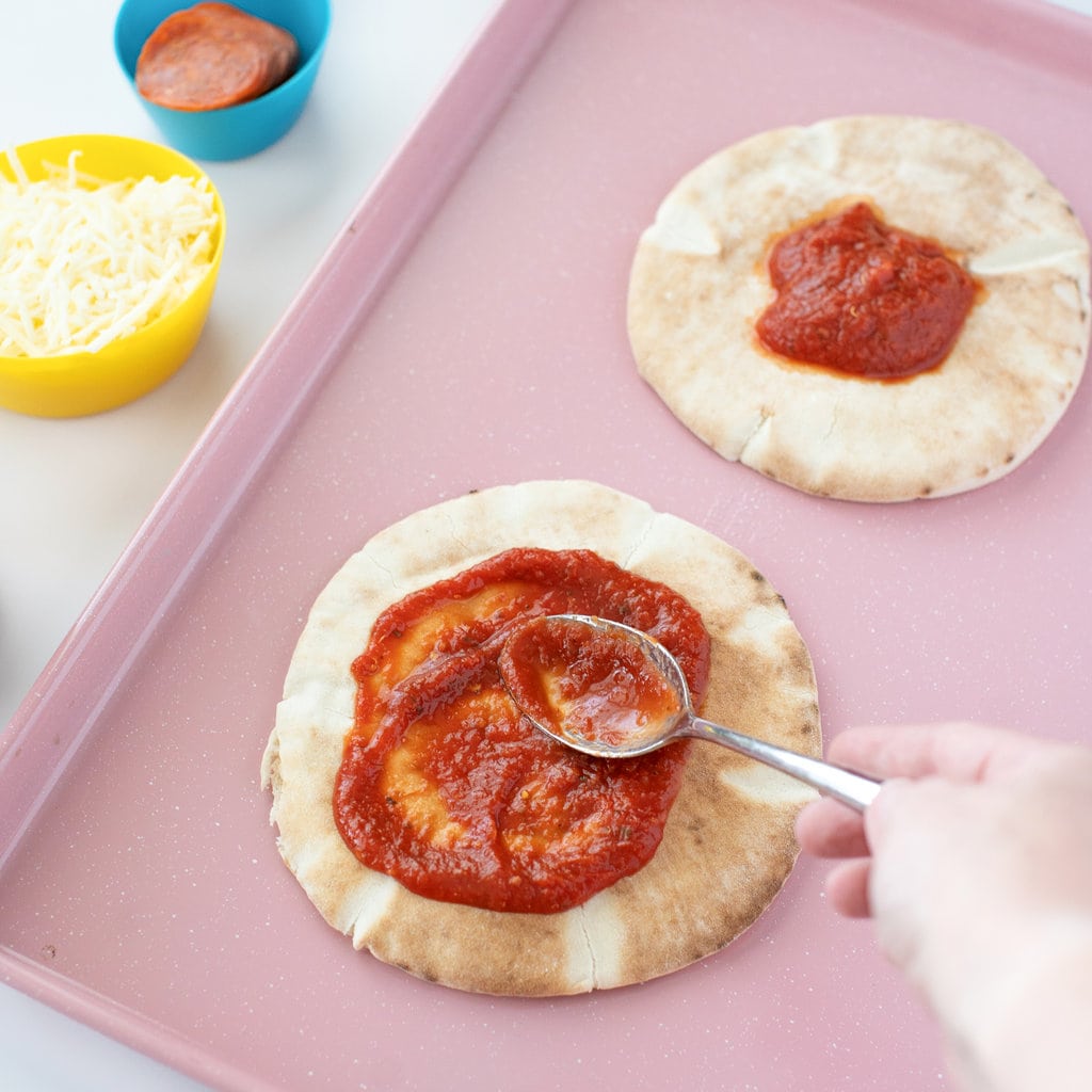 Hand spooning on a pizza sauce to the pita pizza.