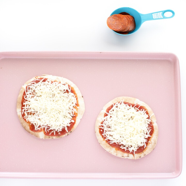 A pink baking sheet with 2 pita pizzas with sauce and cheese.