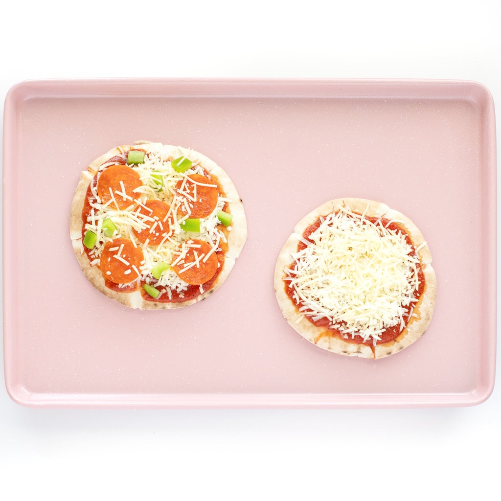A pink baking sheet with 2 pita pizzas with cheese and toppings ready to go into the oven.
