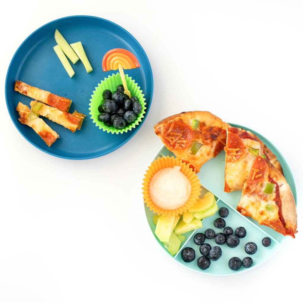 Pita pizza on a teal plate with a side of cucumbers and ranch and blueberries. Another dark blue plate with pita pizza cut into smaller strips for a toddler.