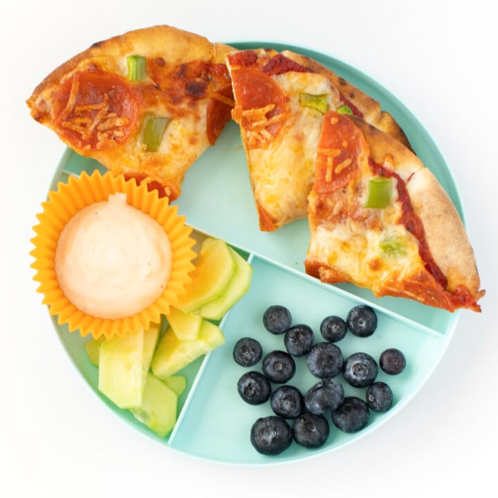 Pita pizza on a teal plate with a side of cucumbers and ranch and blueberries.