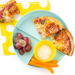A kids teal section plate filled with pita pizza with toppings, a side of ranch and peppers, and cut grapes on a white background with a yellow polkadot napkin.