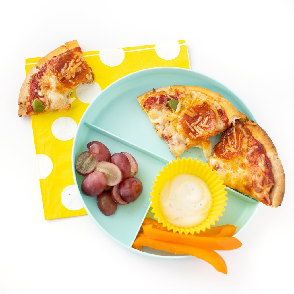 A kids section plate with pita pizza with toppings ranch dressing and a side of grapes, on top of a yellow napkin with part of a pizza with a bite taken out.