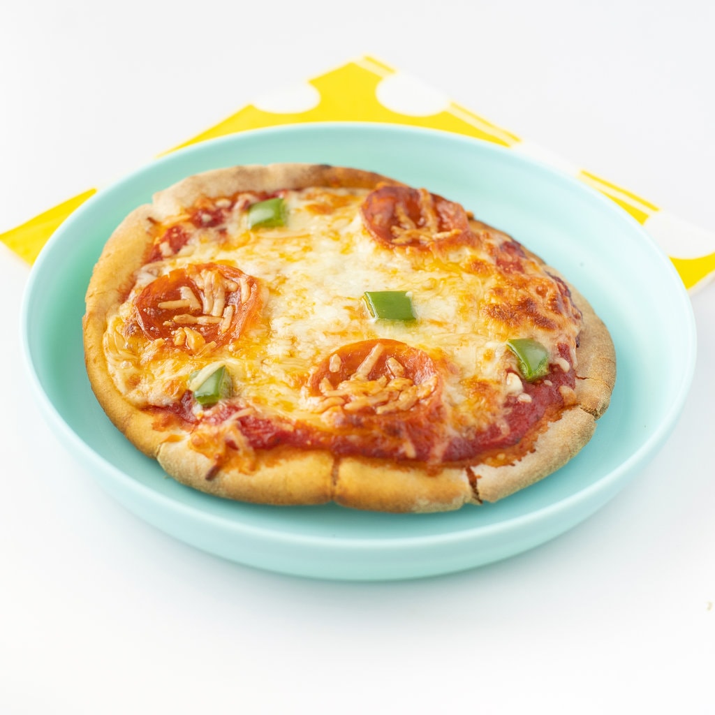 A pita pizza on a teal plate with a yellow napkin against a white background. 