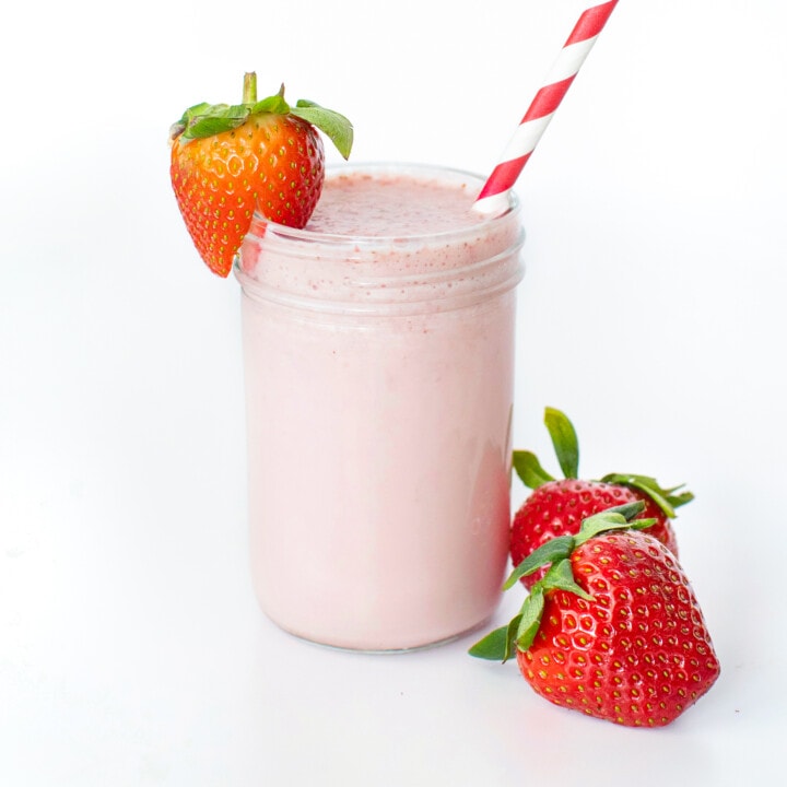 Clear glass filled with a pink strawberry milk with a strawberry on the lip and a striped straw.