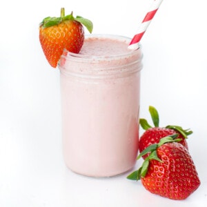 Clear glass filled with a pink strawberry milk with a strawberry on the lip and a striped straw.