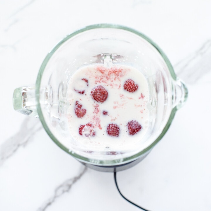 Clear blender on white marble counter with ingredients for strawberry milk.
