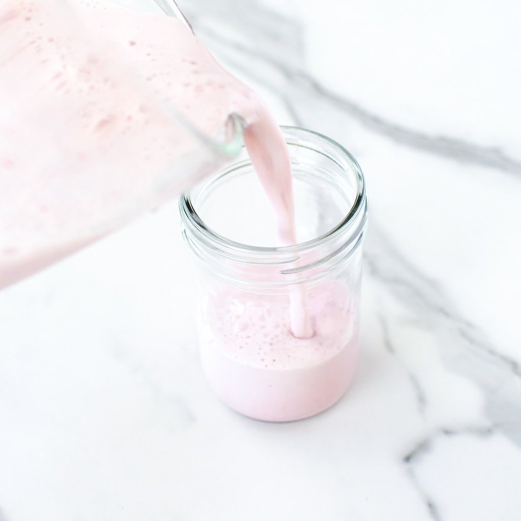 Clear blender pouring strawberry milk inside a clear class on a marble white counter. 