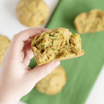 Small hand holding a muffin over a table with muffins and a green napkin.