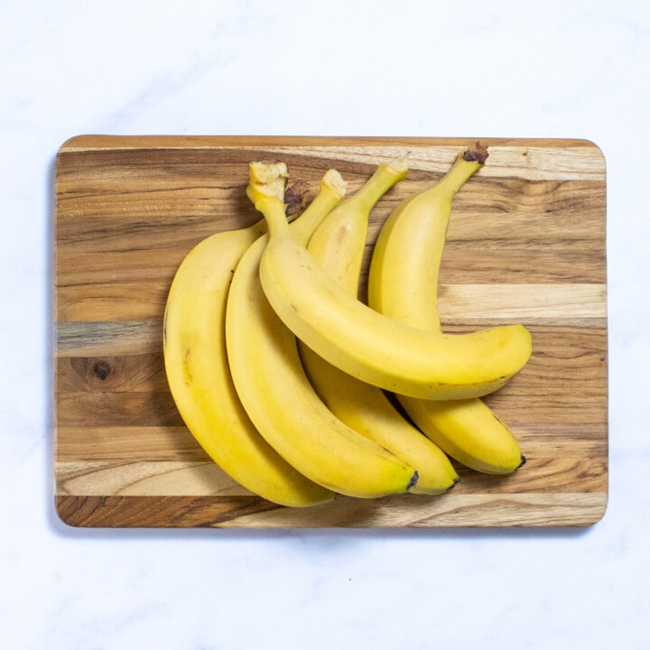 Wooden cutting board on a marble countertop with bananas piled on top.