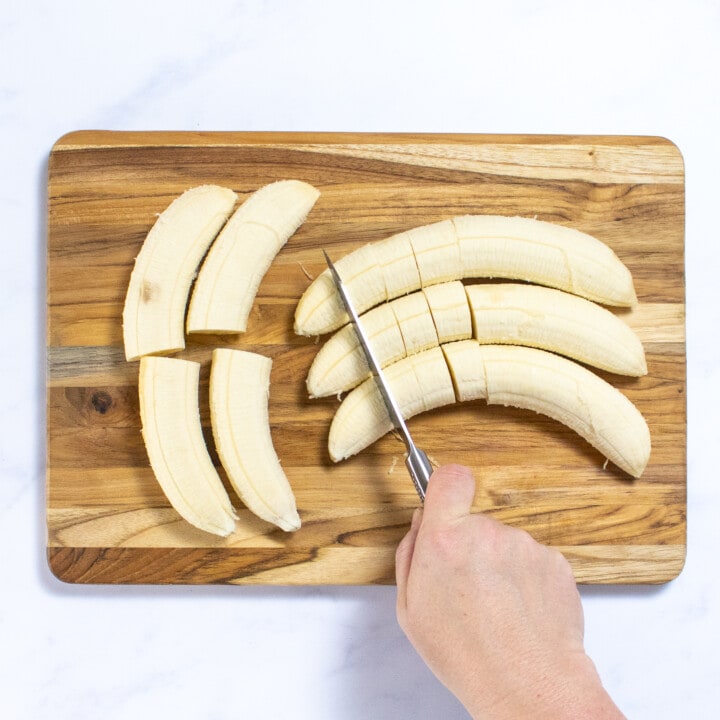 A hand holding a knife cutting bananas into slices on a wood cutting board.
