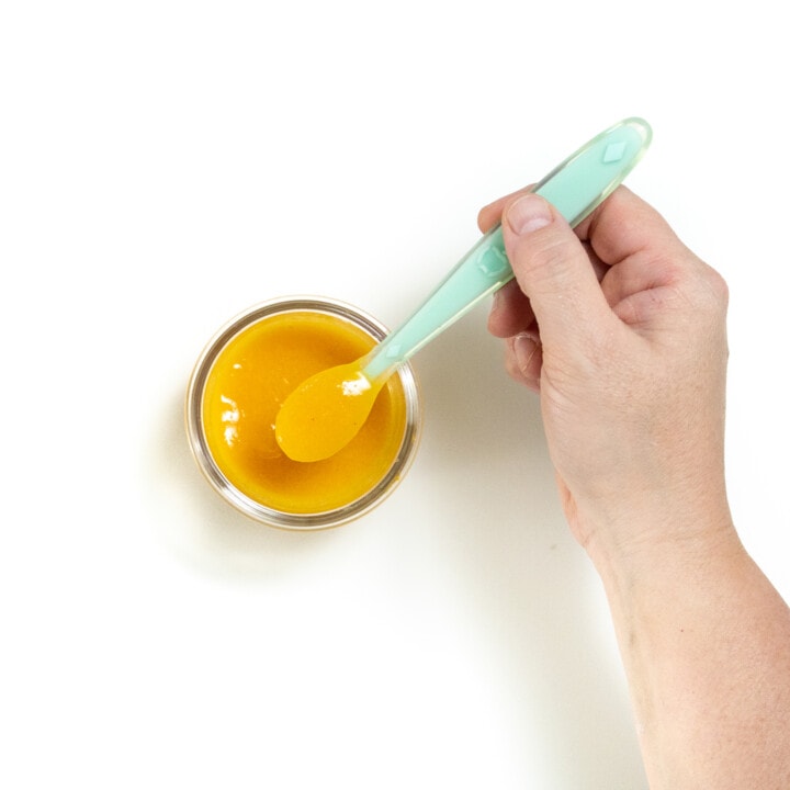 A hand holding a baby spoon with apricot baby food purée over a glass jar against a white background.