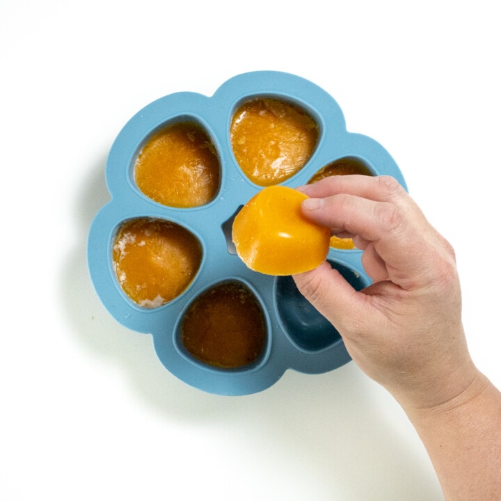 a Hand holding and frozen cube of apricot baby food purée over a blue freezer tray background.