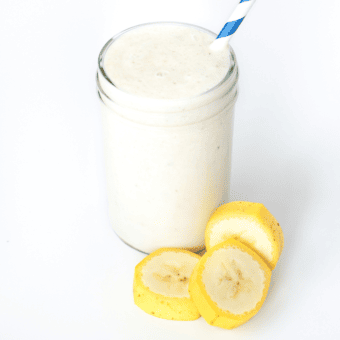 A glass of banana milk against a white background with a blue stripe straw and rounds of banana next to it.