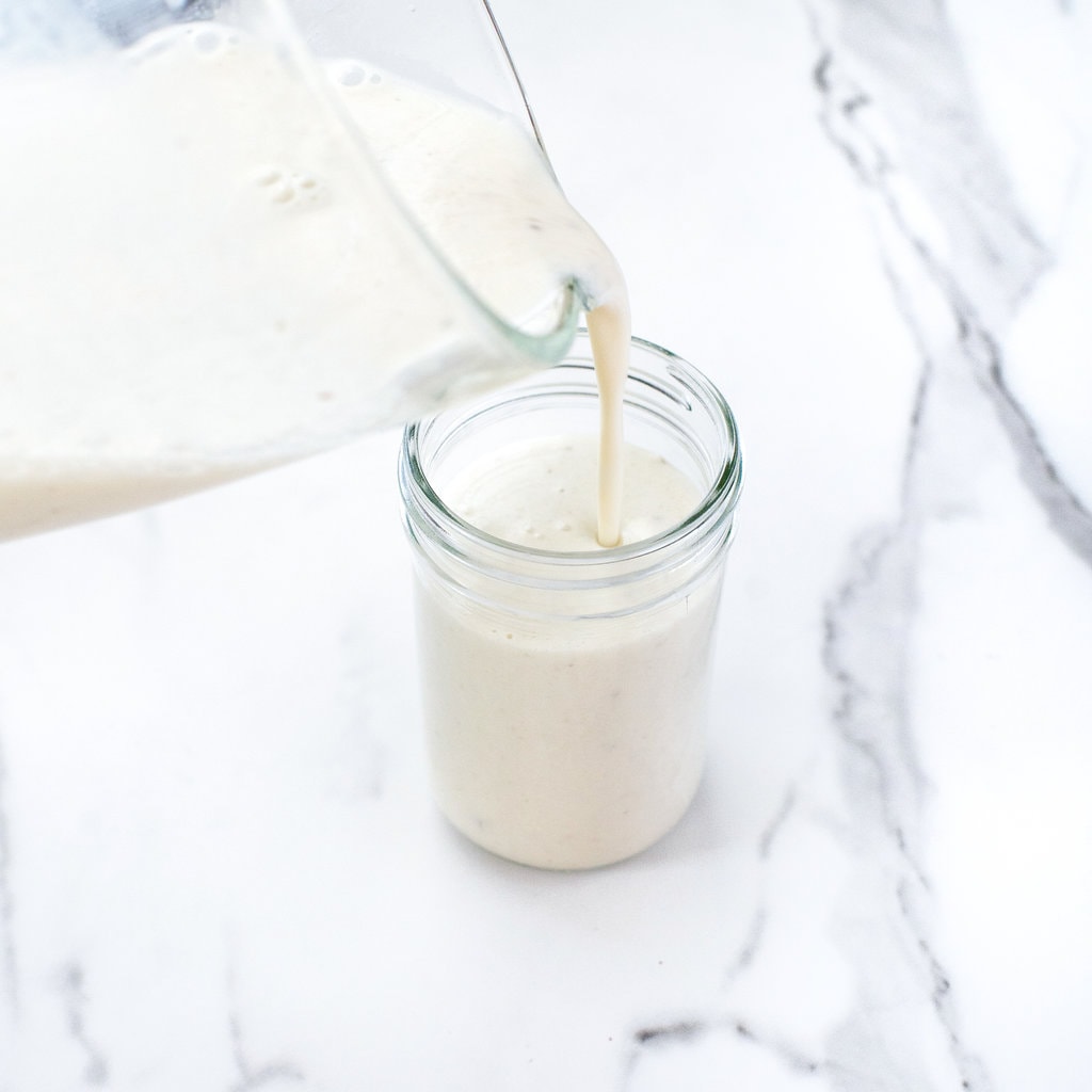 A clear blender pouring banana milk into a clear cup on a white marble background.