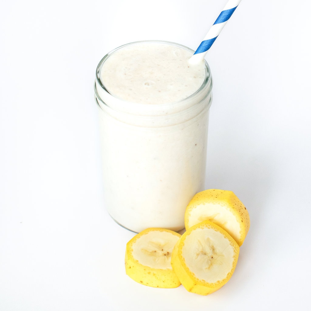 A glass of banana milk against a white background with a blue stripe straw and rounds of banana next to it.