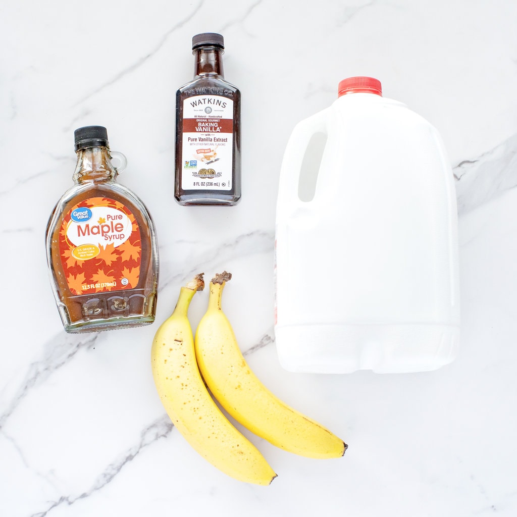 Ingredients needed for banana milk – milk, bananas, vanilla extract, and maple syrup, on a marble countertop.