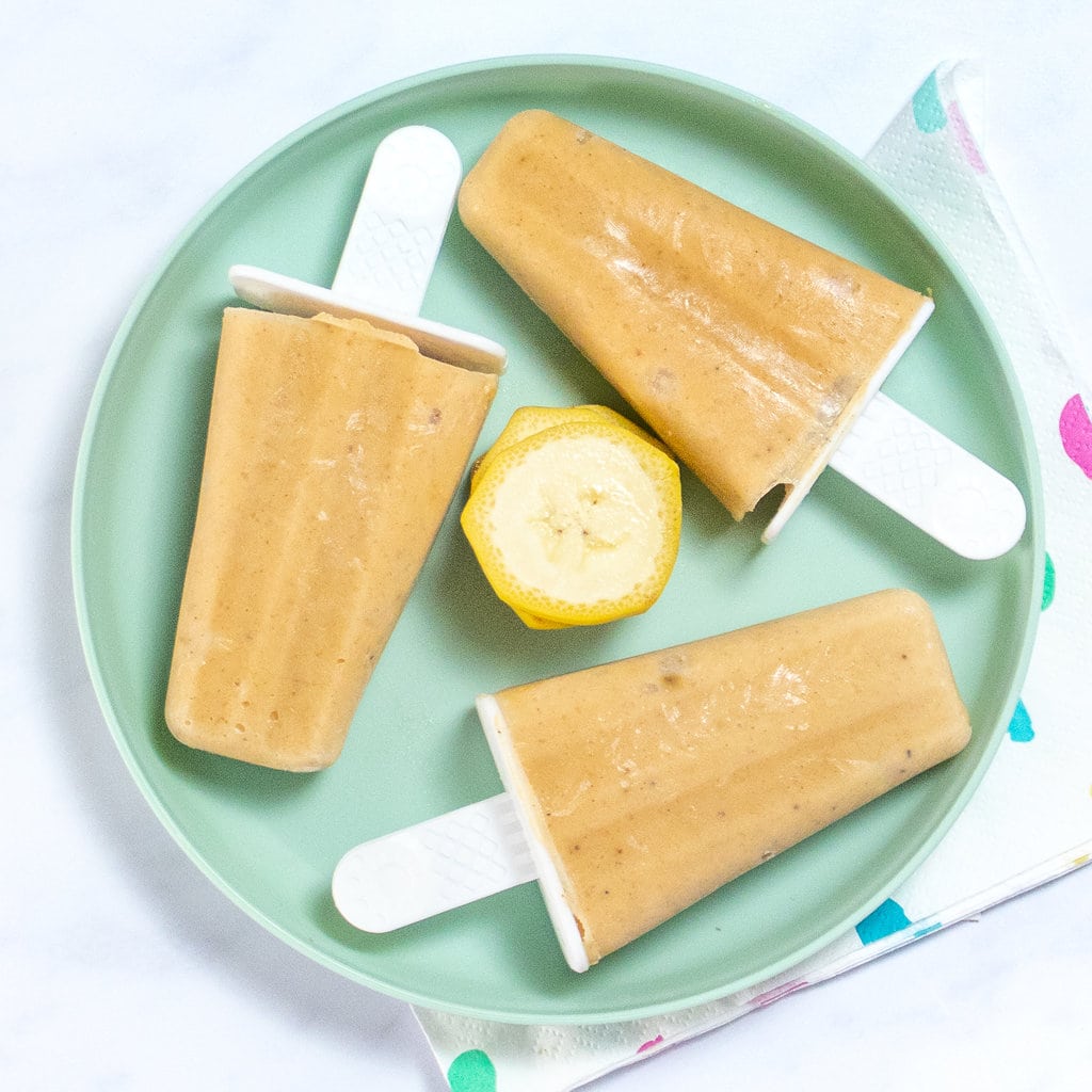 A kids teal plate with banana popsicles and slices of bananas against a white marble counter.