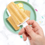 A hand holding up a popsicle over a plate of banana popsicles and a colorful napkin. The teal plate is on a colorful napkin sitting on my white marble kitchen counter.
