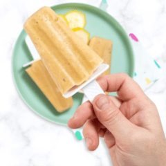 A hand holding up a popsicle over a plate of banana popsicles and a colorful napkin. The teal plate is on a colorful napkin sitting on my white marble kitchen counter.