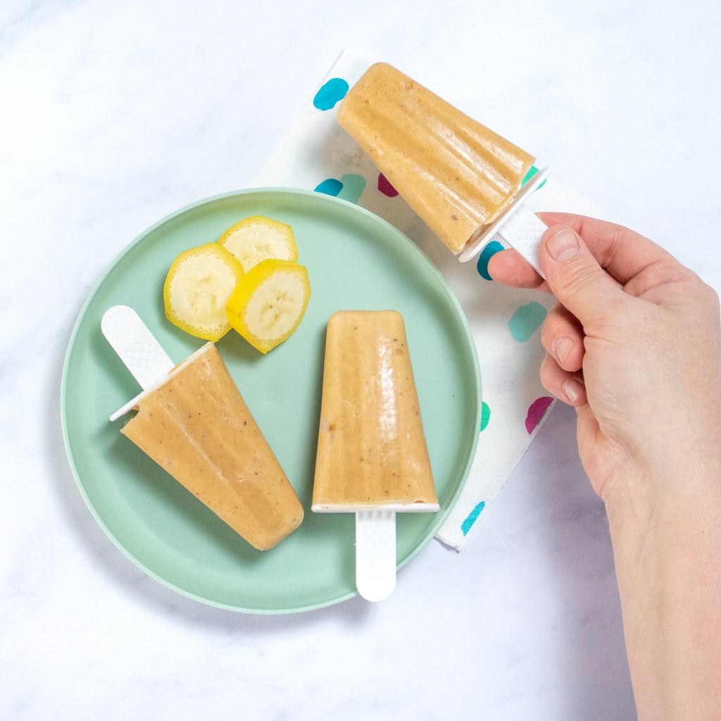 Hand holding a banana popsicle over a colorful napkin next to a plate with more popsicles and a few slices of banana.