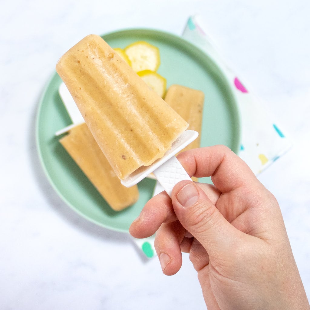 A hand holding up a popsicle over a plate of banana popsicles and a colorful napkin.