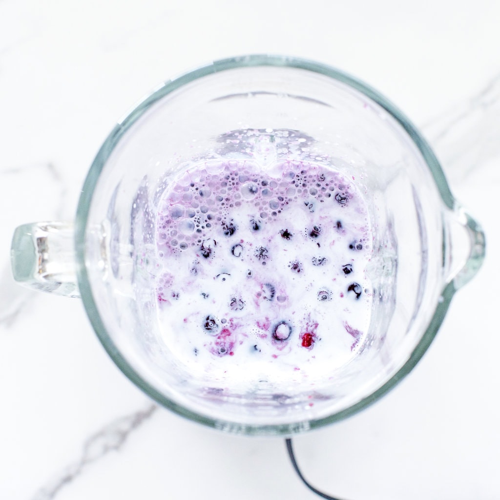 A clear blender against a marble countertop with blueberries, milk, maple syrup, and vanilla.