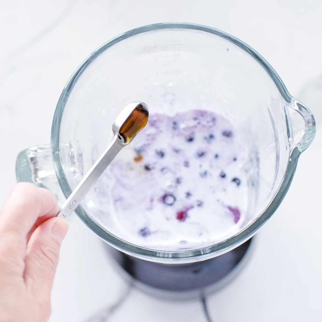 Hand pouring syrup into a blender full of blueberries, and milk.