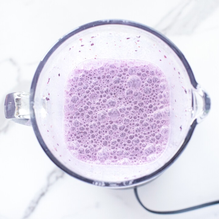 A clear glass blender with blueberry milk with bubbles on top.