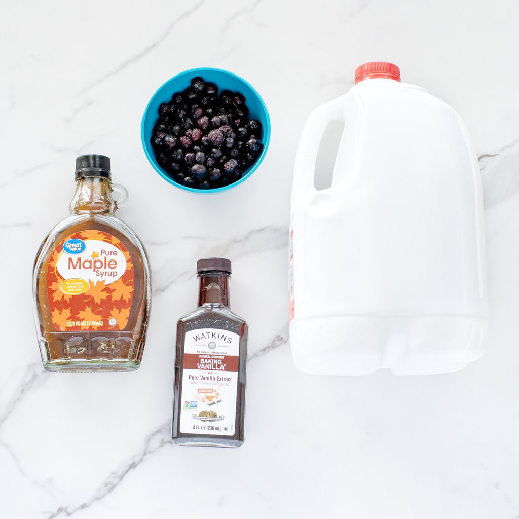 Ingredients needed to make blueberry milk spread on the counter – milk, vanilla, maple syrup and blueberries.