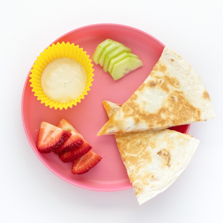 A pink kids plate with wedges of pizza quesadilla sliced grapes and cucumber with ranch dipping sauce.