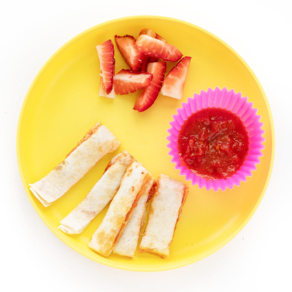 A pizza quesadilla cut for a toddler in long strips with a cup of marinara sauce and chopped strawberries.