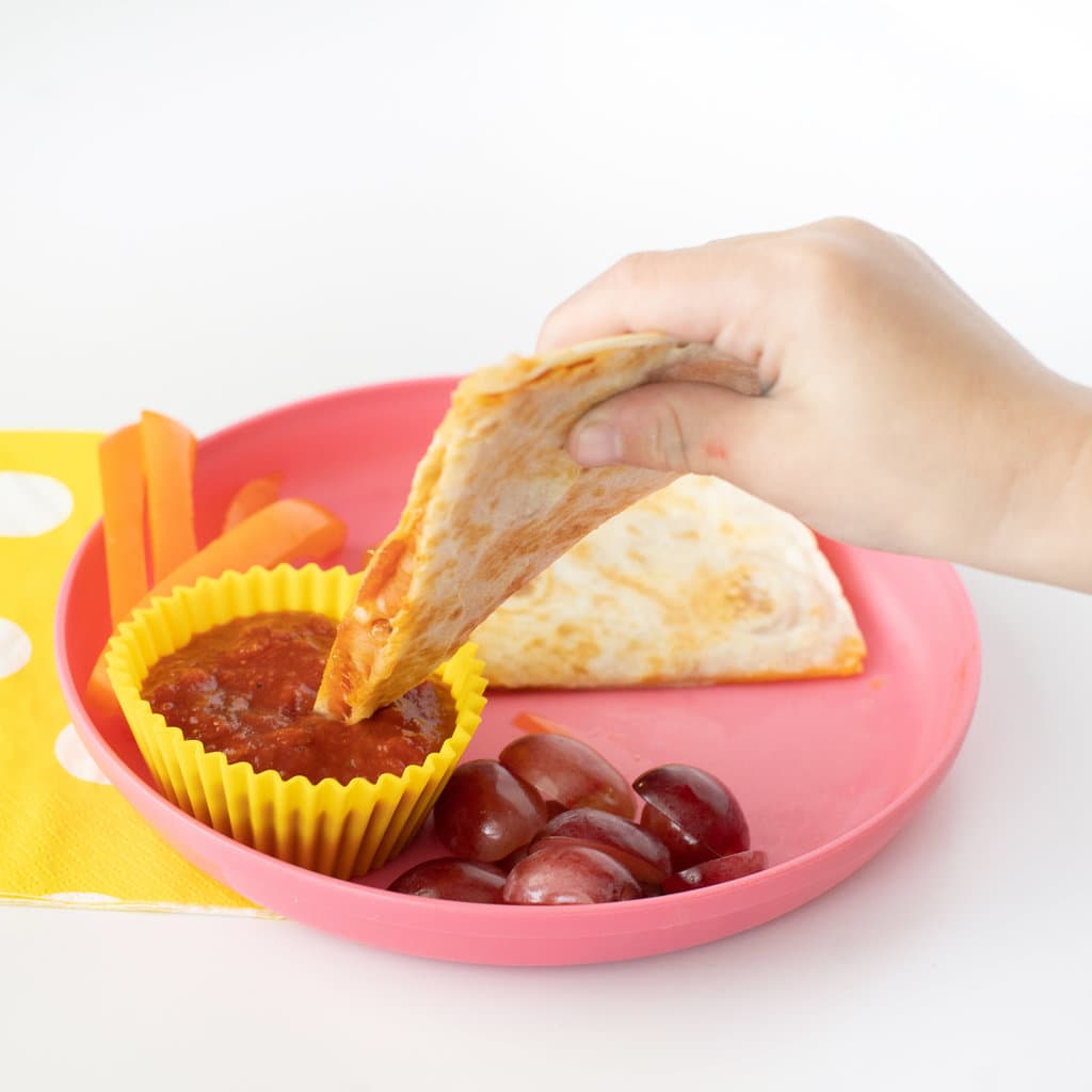 A kids hand holding a pizza quesadilla dipping it in marinara sauce with a pink plate against a white background.